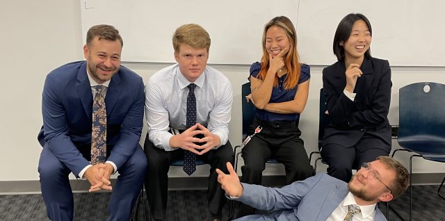 4 graduate students in suits in chairs and one graduate student in a suit leaning on the floor