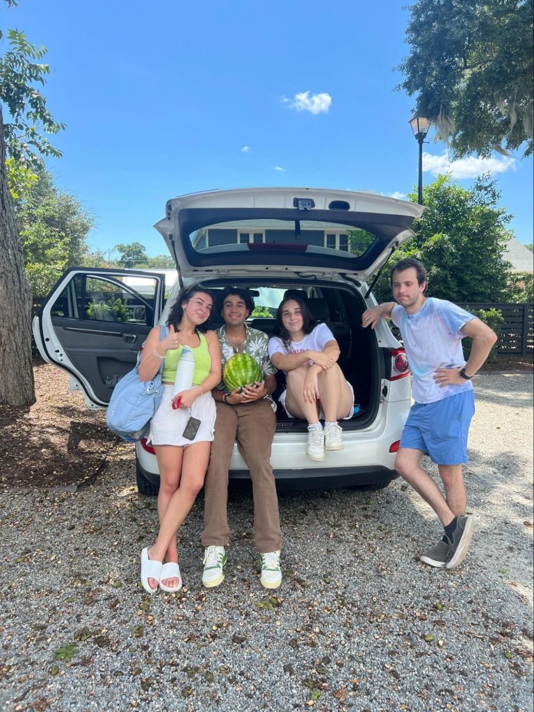 Two people sitting in the back of a car and two people leaning agains the car. One of the people sitting is holding a watermelon. 